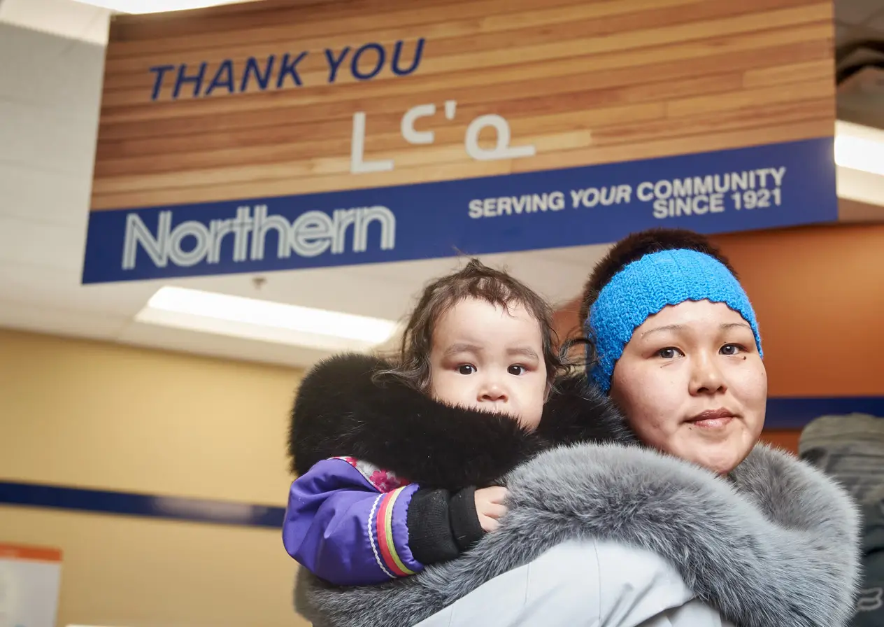 mother in traditional amauti with a baby, standing in front of the Thank You sign at Northern Store