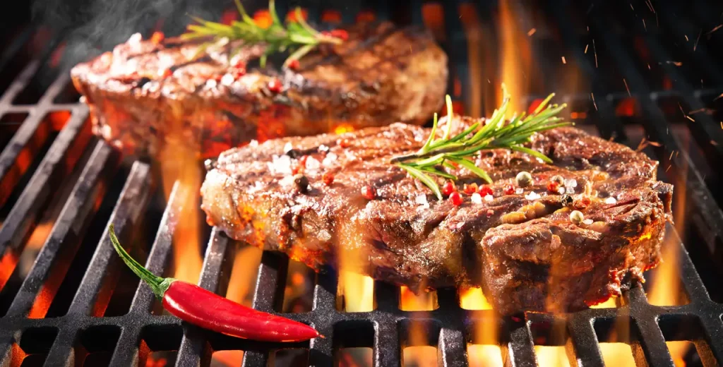 A couple of Beef burgers cooking on a BBQ with a red pepper beside them